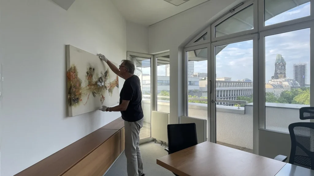 Mitarbeiter von ARTprotect hängt ein Originalgemälde auf der Dachterrasse eines Berliner Büros mit Stadtblick auf